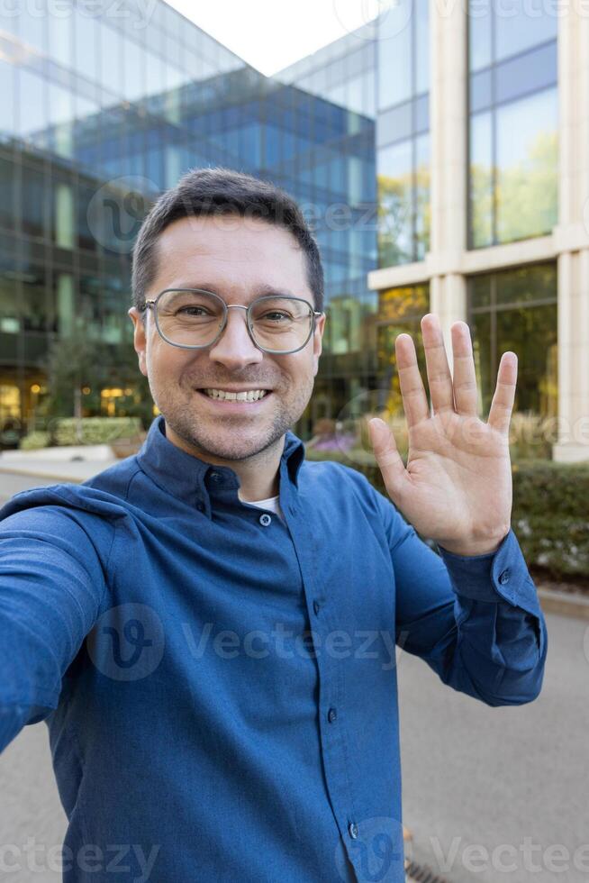 A man waves at the camera, smiling broadly, with a modern building in the background. The man wears glasses and a blue shirt. photo