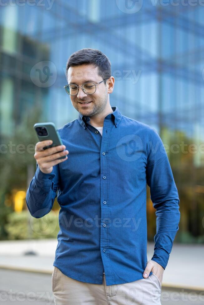 A man in glasses is using a smartphone outside in front of a modern office building, he seems pleased. photo