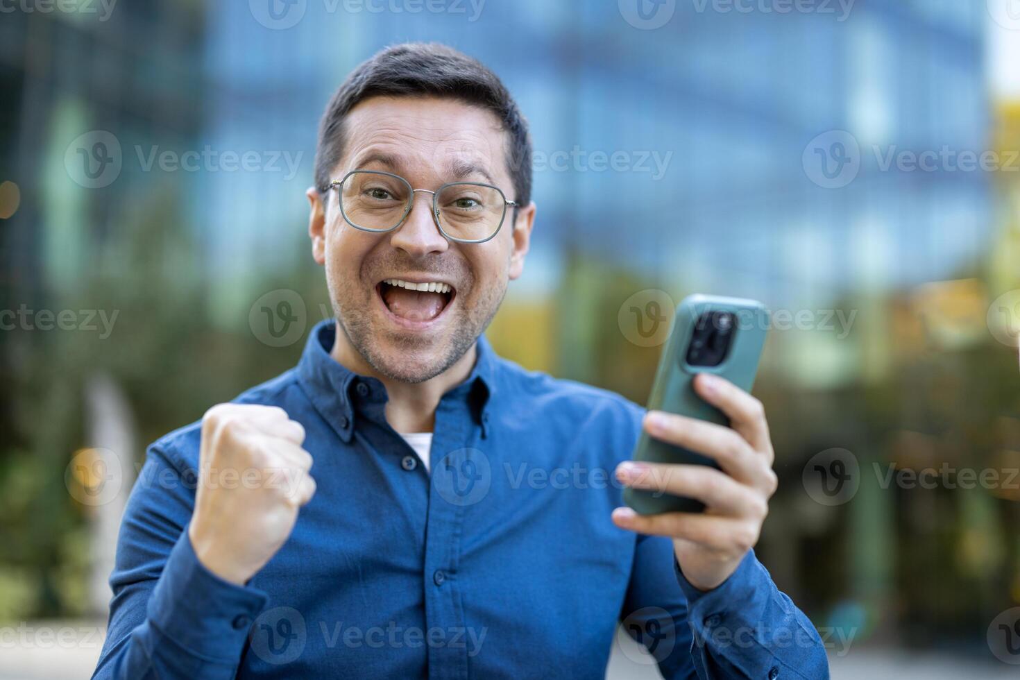 A man joyfully reacts to something he sees on his phone, fist clenched in celebration while outside. photo