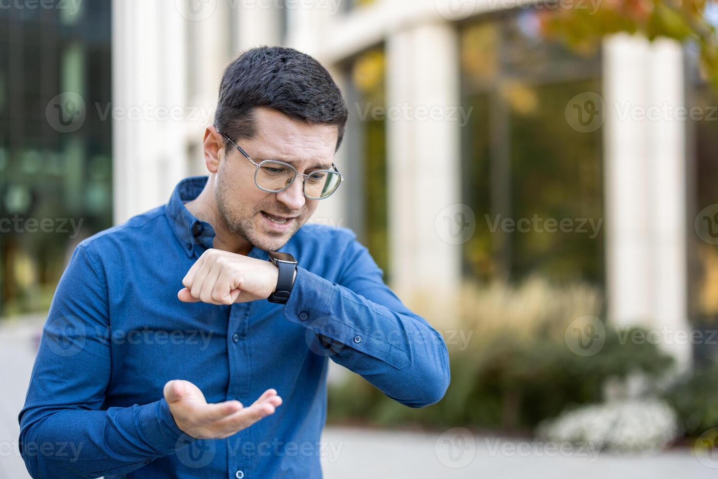 A man wearing glasses uses a smartwatch to talk using an app on the street in front of a building photo