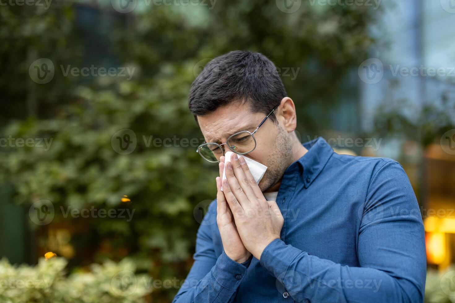 A man in glasses outside, blowing his nose with a tissue, suggesting allergies or illness. photo