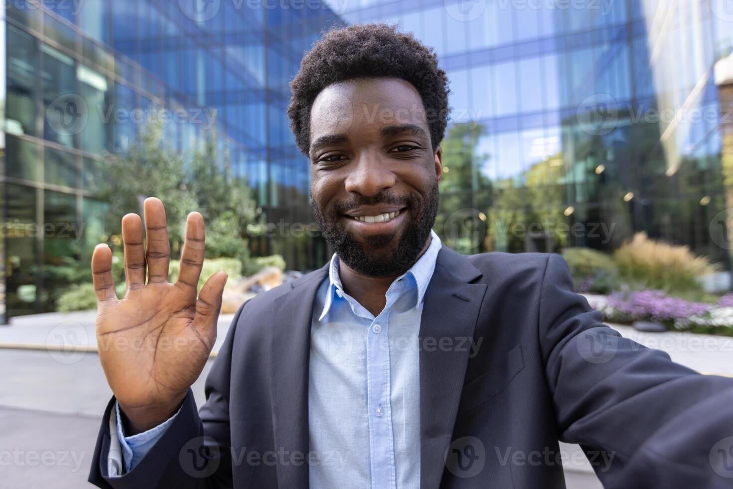 An attractive man in a business suit is waving in front of a modern building. Smiling directly at the camera, outdoors. photo