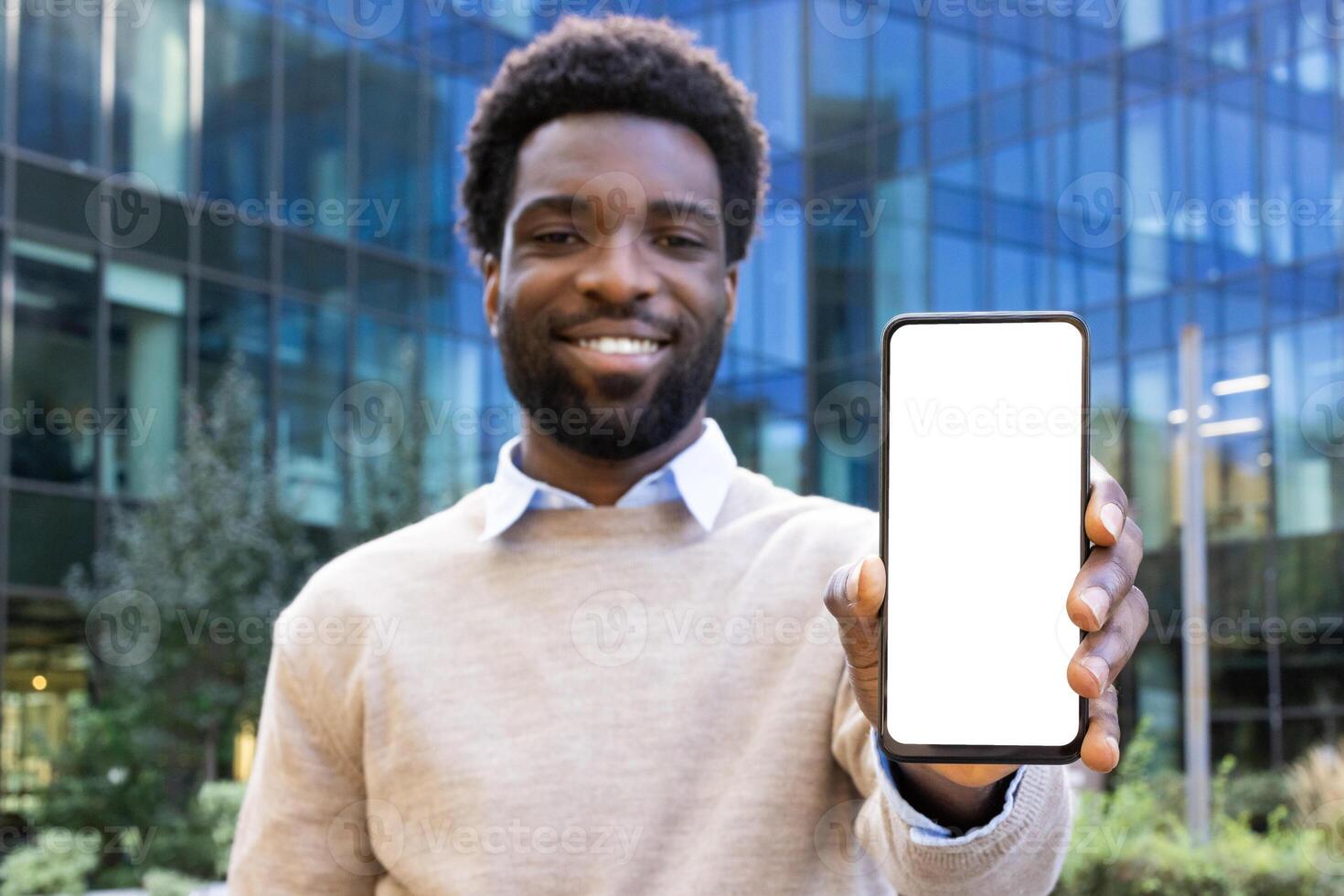 Smiling man holding up a phone with a blank screen in front of a modern glass building. The photo is taken in daylight.
