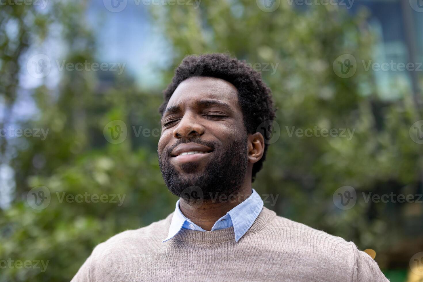 A happy black man with closed eyes enjoys the fresh air, expressing serenity and joy in a park setting, near a building. photo