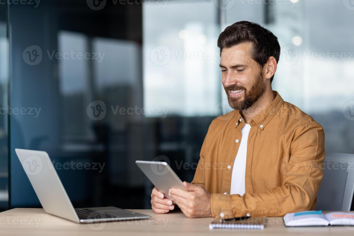 A smiling man is sitting at a desk, holding a tablet, with a laptop nearby, indoors at the office. photo