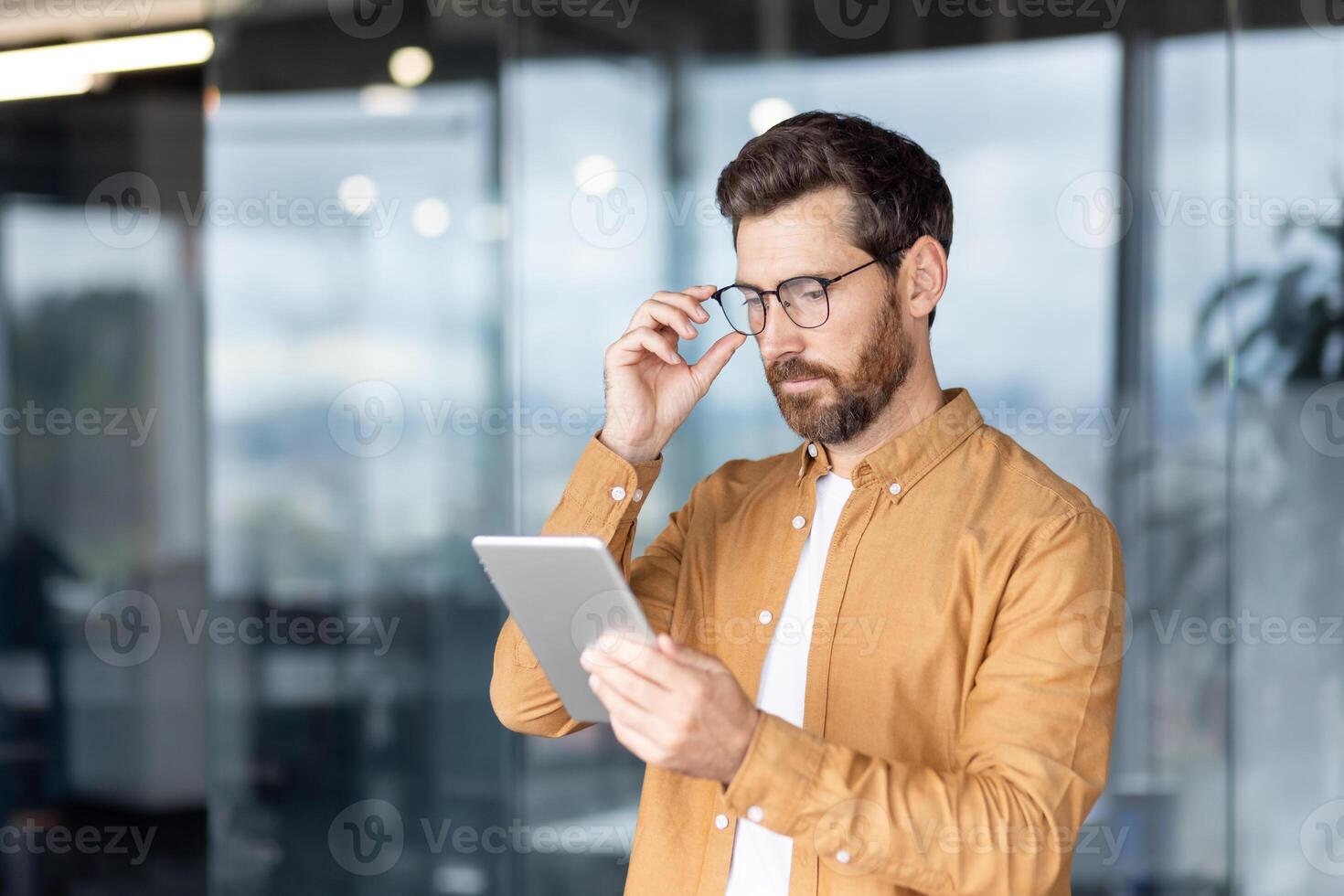 A focused businessman reviews a tablet while adjusting his glasses in a modern office setting, suggesting productivity and technology. photo