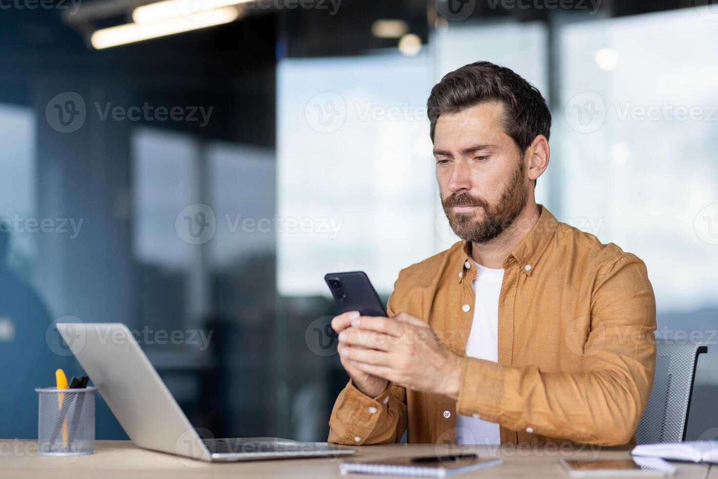 A focused man uses his smartphone while sitting at a desk in an office setting. Laptop and pen holder on the table. photo