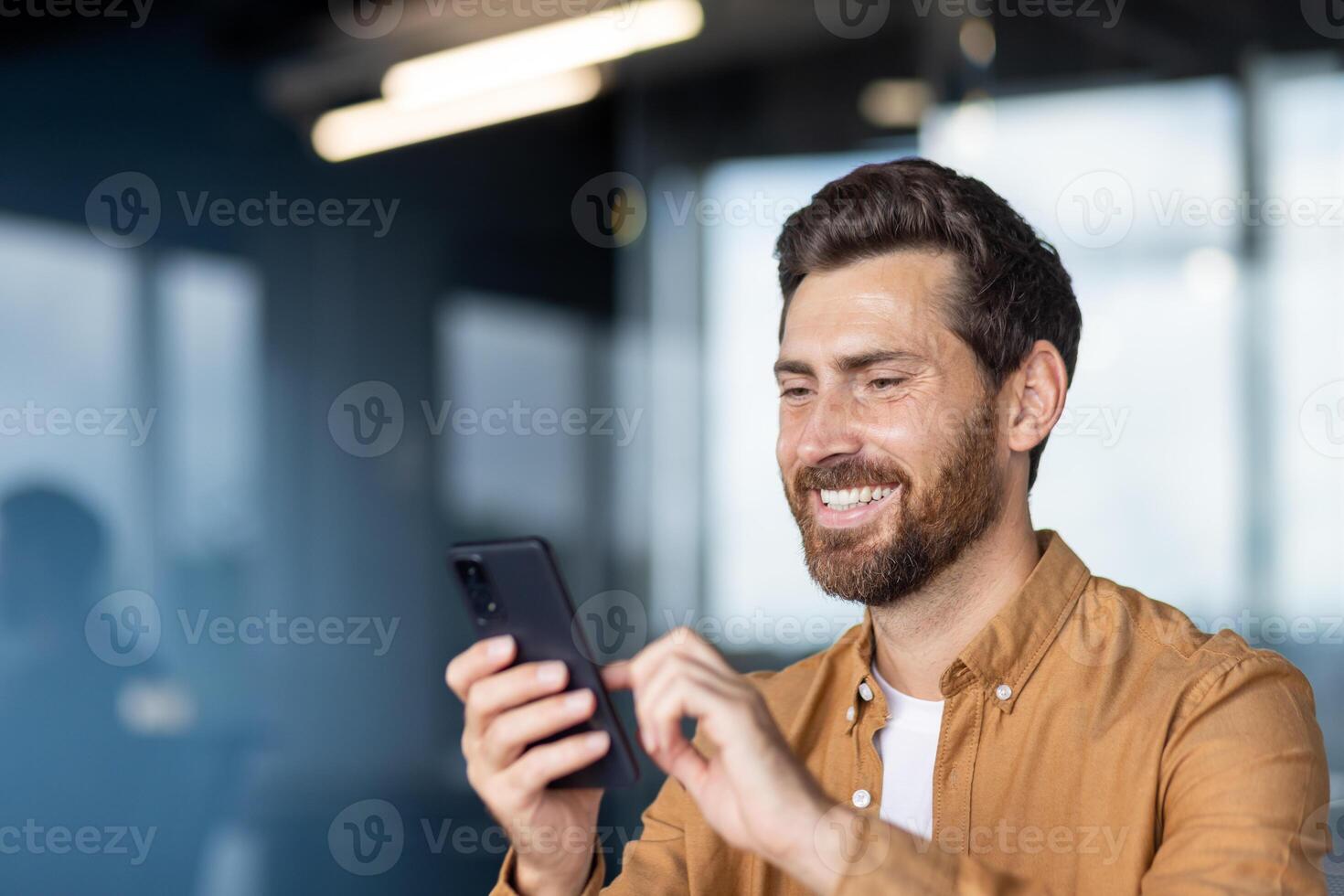A smiling man in a button-down shirt looks at his phone, likely texting or browsing in a modern office setting. photo
