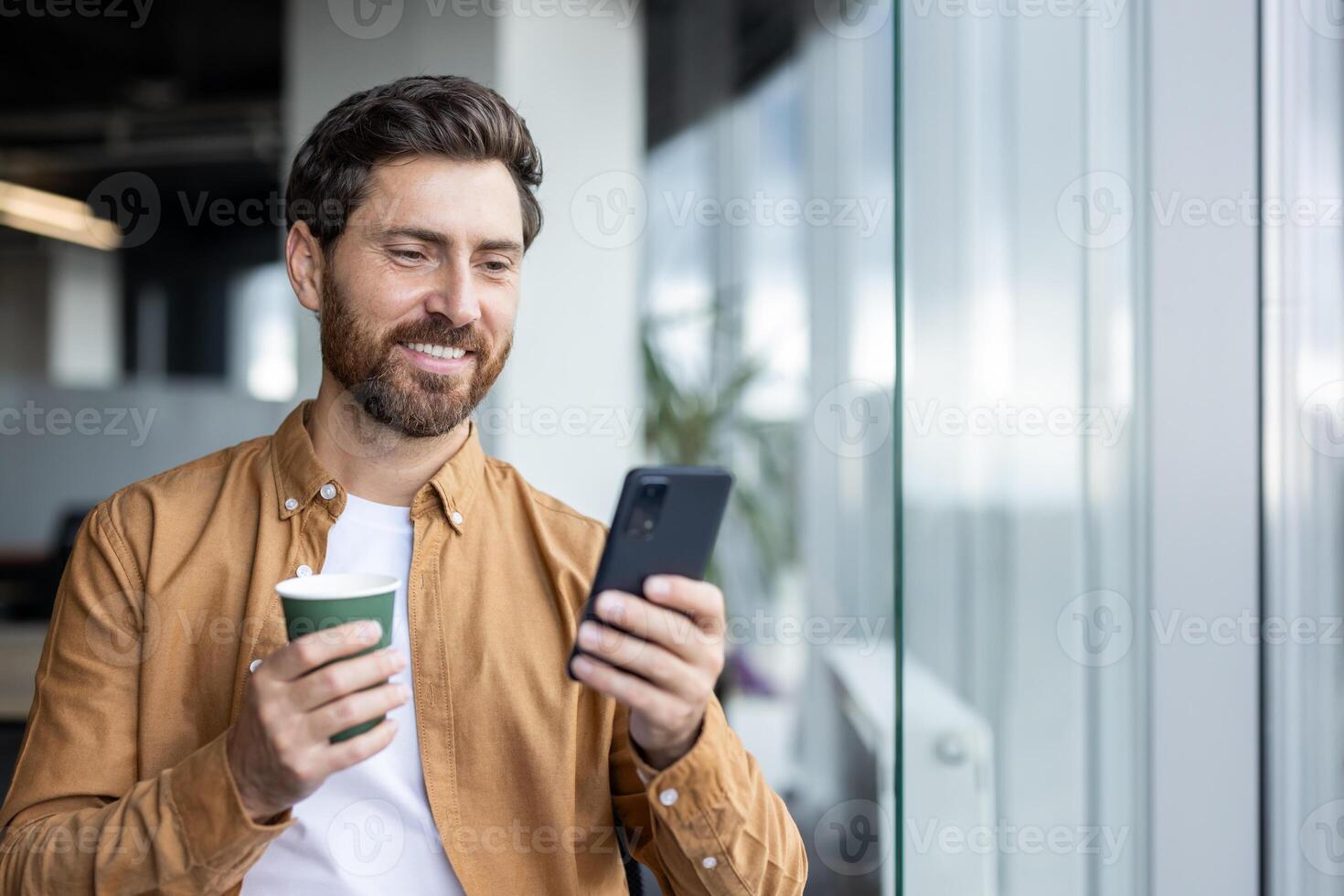 Successful male businessman inside office standing by window with phone in hands and cup of hot drink. Businessman browsing social media and typing text message. photo