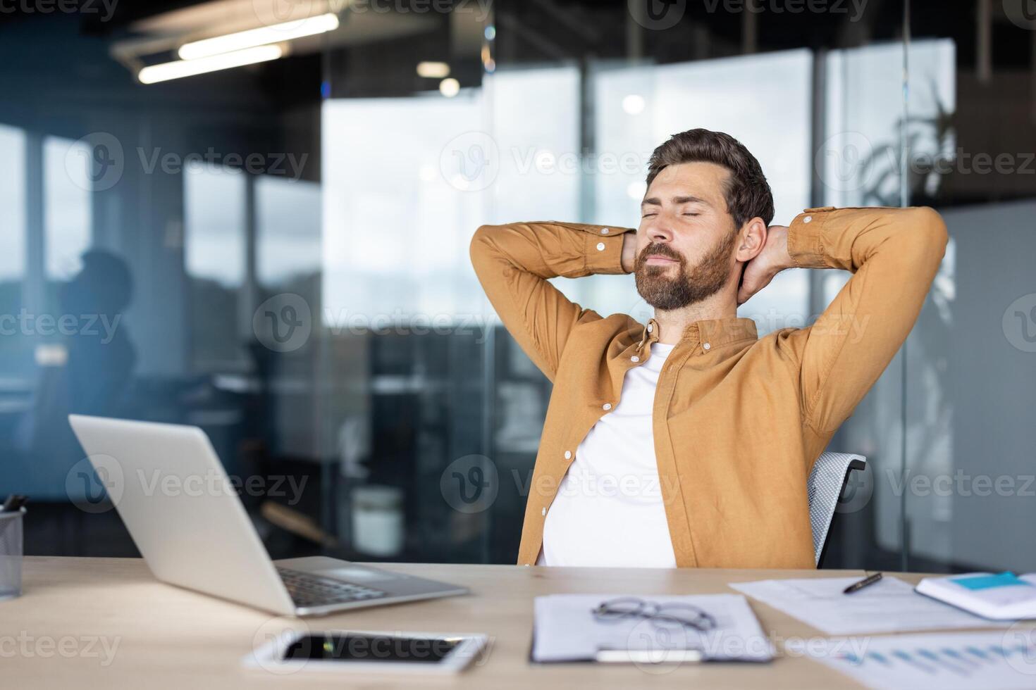 A relaxed businessman with closed eyes leans back in his office chair, hands behind his head, laptop on the desk in front of him. photo