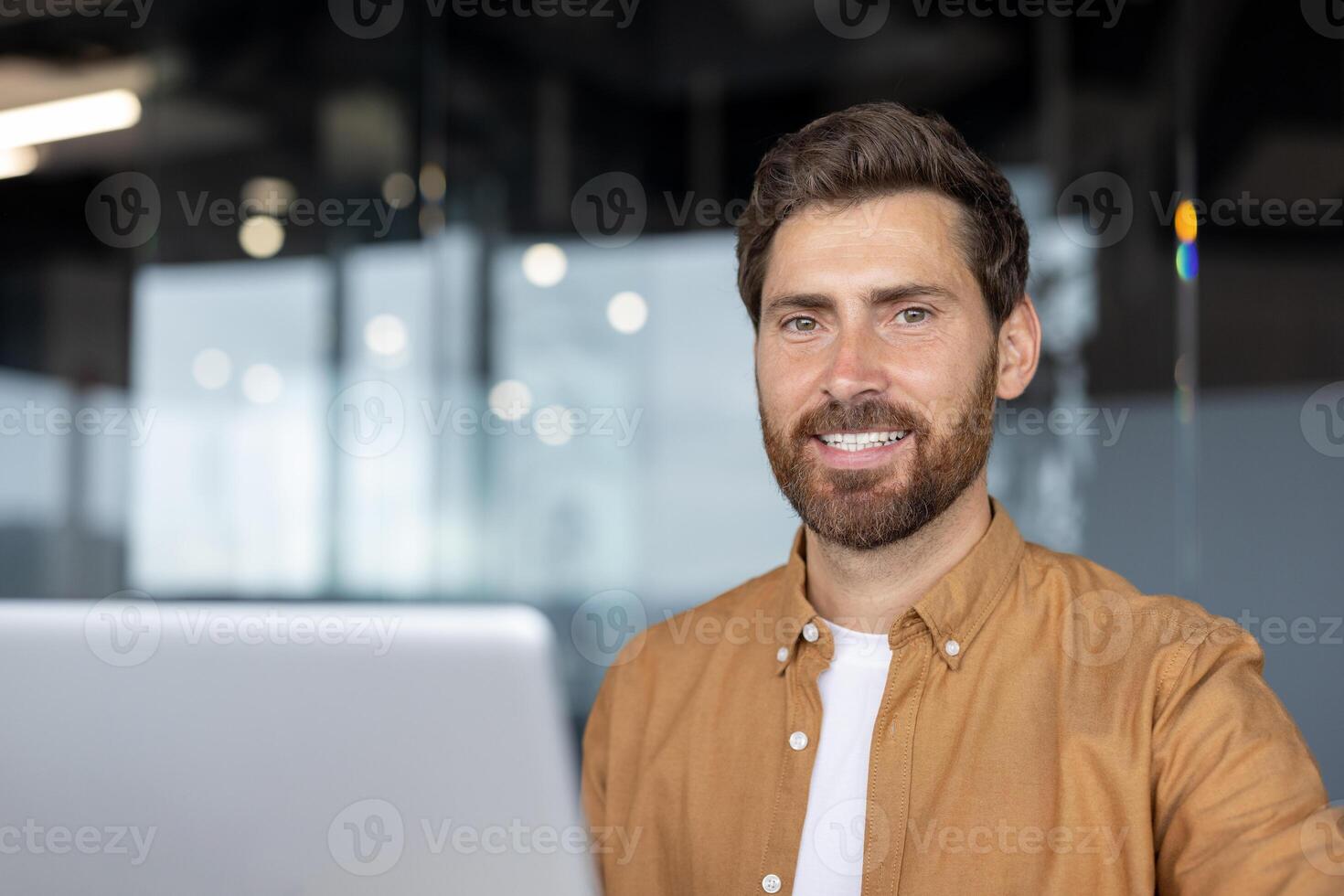 Mature businessman smiling in office with laptop. Casual shirt conveys relaxed yet professional demeanor. Confident expression suggests positive attitude and success in work environment. photo