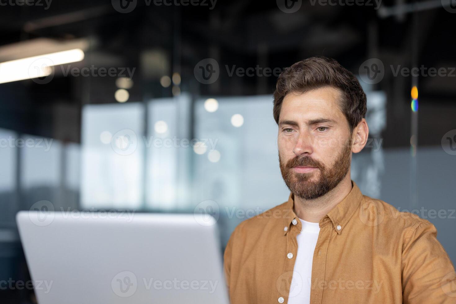 Man with beard using laptop in office. Reflects concentration, focus, productivity in modern workspace. Casual wear suggests informal but professional setting. photo