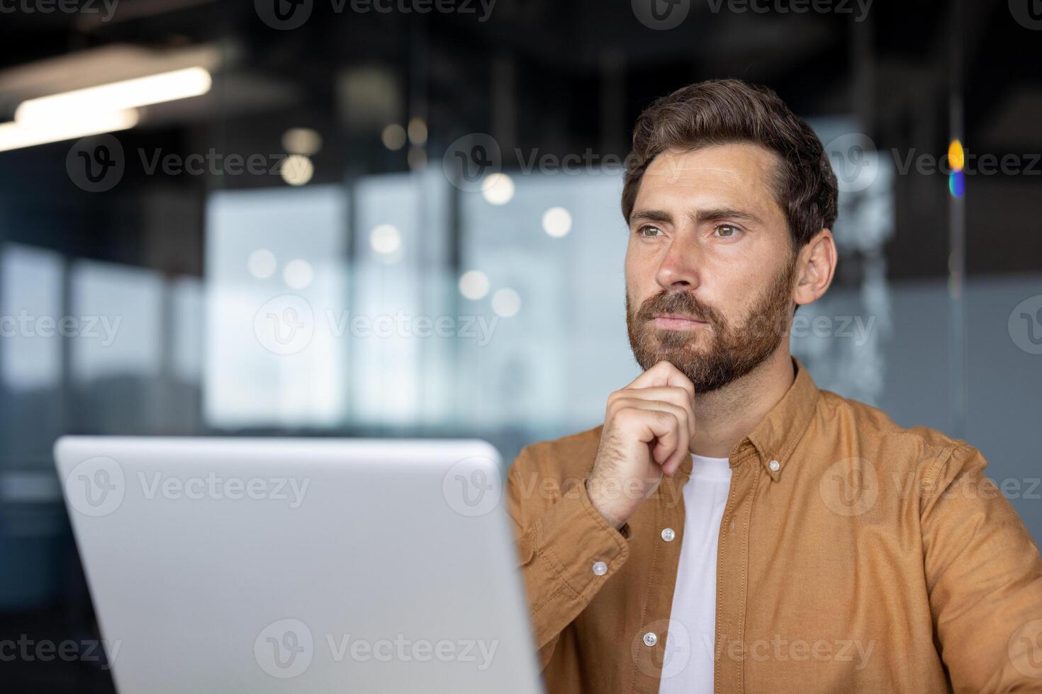 Man in casual shirt thinking while using laptop in modern office setting, reflecting on work ideas. Natural light filters through glass walls, creating focused atmosphere. photo