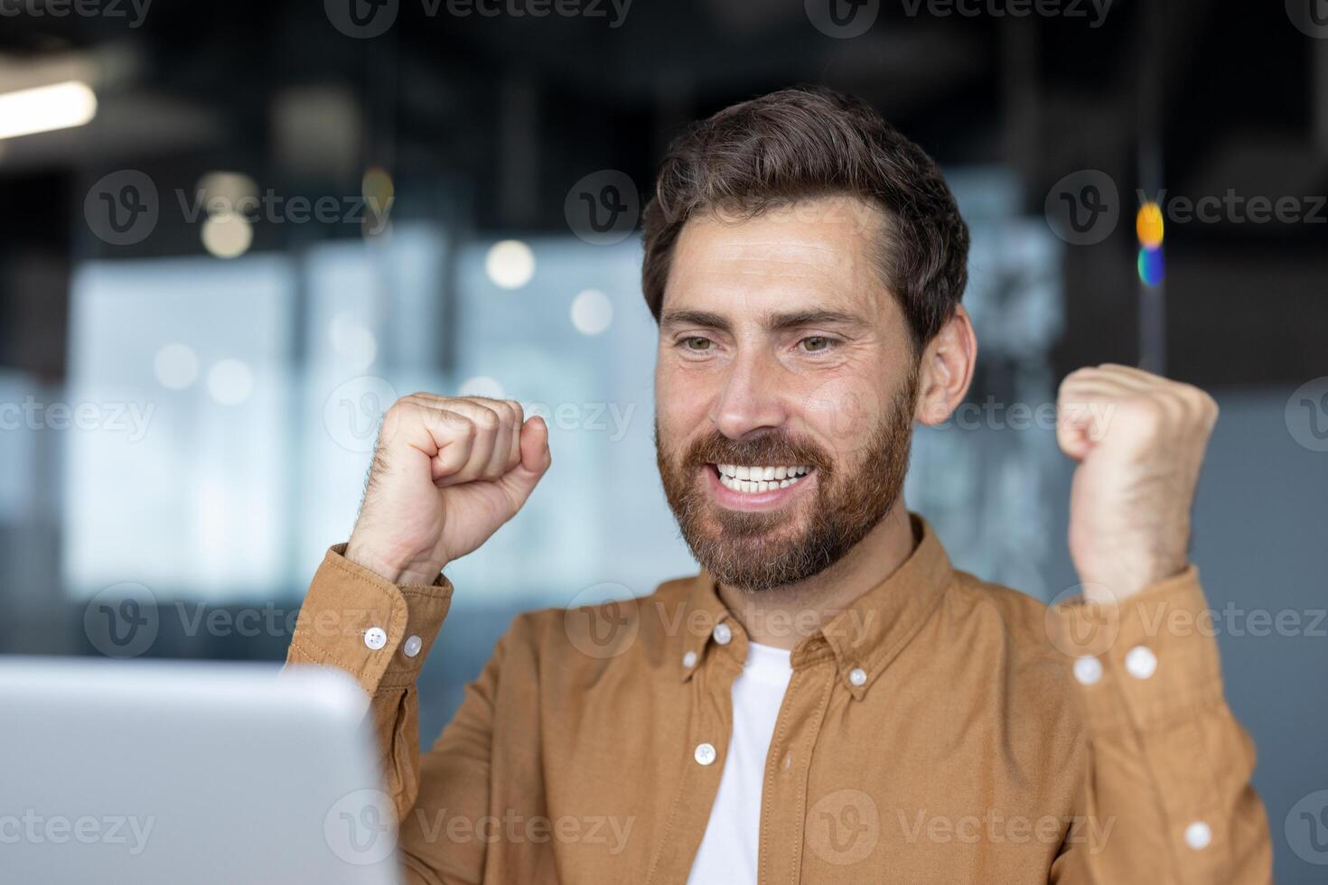 Happy man showing excitement in office, celebrating work success while viewing computer screen. Joyful expression, triumphant fists showcase motivation, achievement, positive attitude. photo