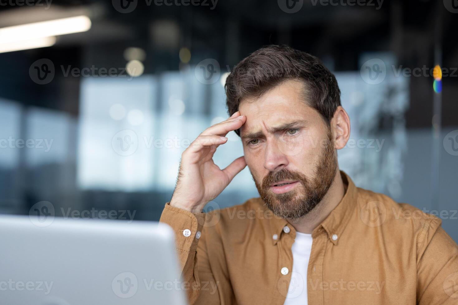 Man in office setting deeply focused on laptop, looking thoughtful, indicating problem-solving. Office environment suggests professional setting with colleagues and conversations unfolding. photo