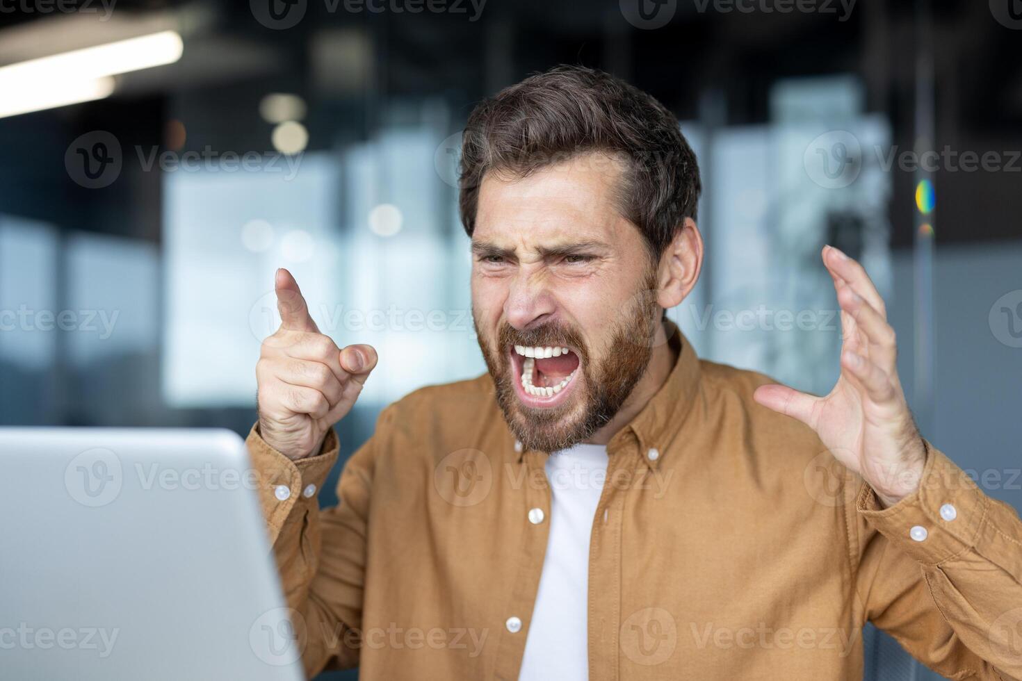 Frustrated businessman yelling at laptop during call. Intense anger and stress evident in facial expressions. Office setting hints at workplace conflict, emotion, frustration, and tension. photo
