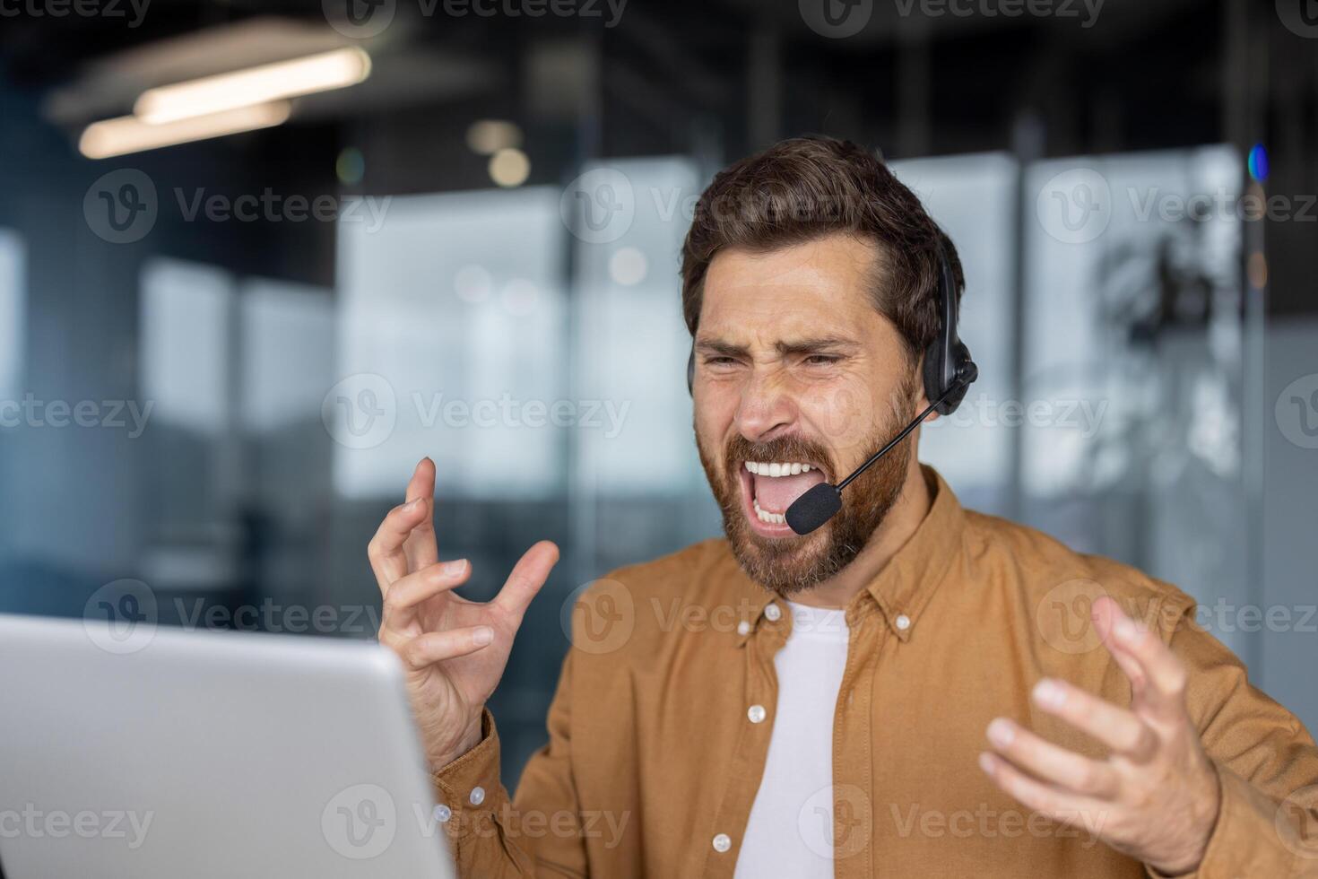 Businessman shows frustration with tech issues during work. Wearing headset, interacting with laptop. Challenges in professional environment, emotion of stress evident in expression. photo