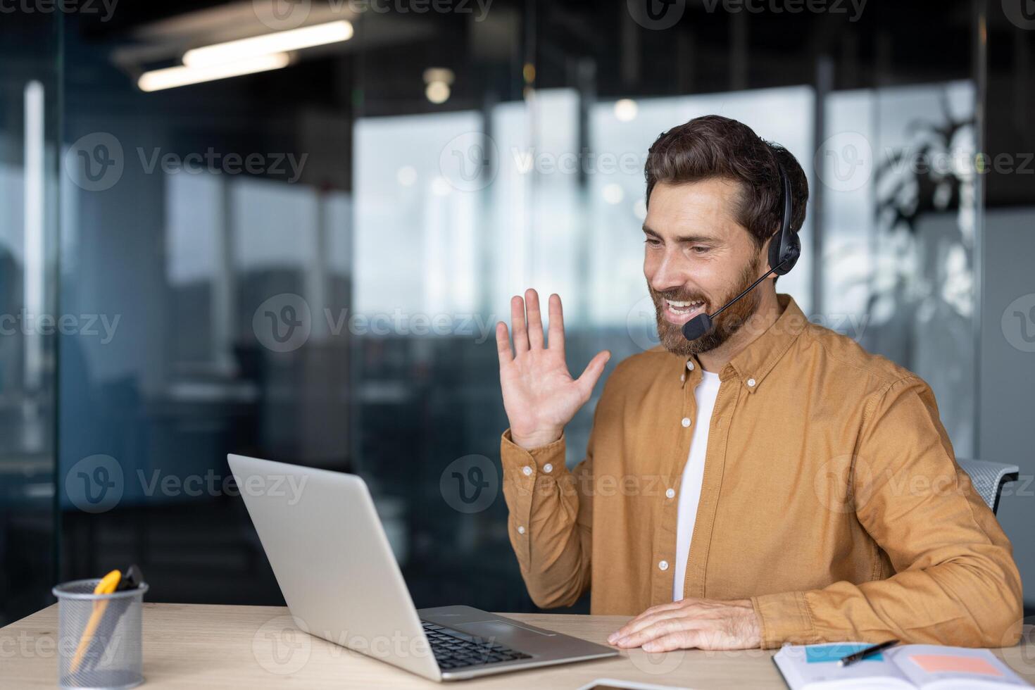 Smiling man in brown shirt using laptop headset for call in modern office setting. Engaging in virtual meeting with colleagues, demonstrating innovation communication technology. photo