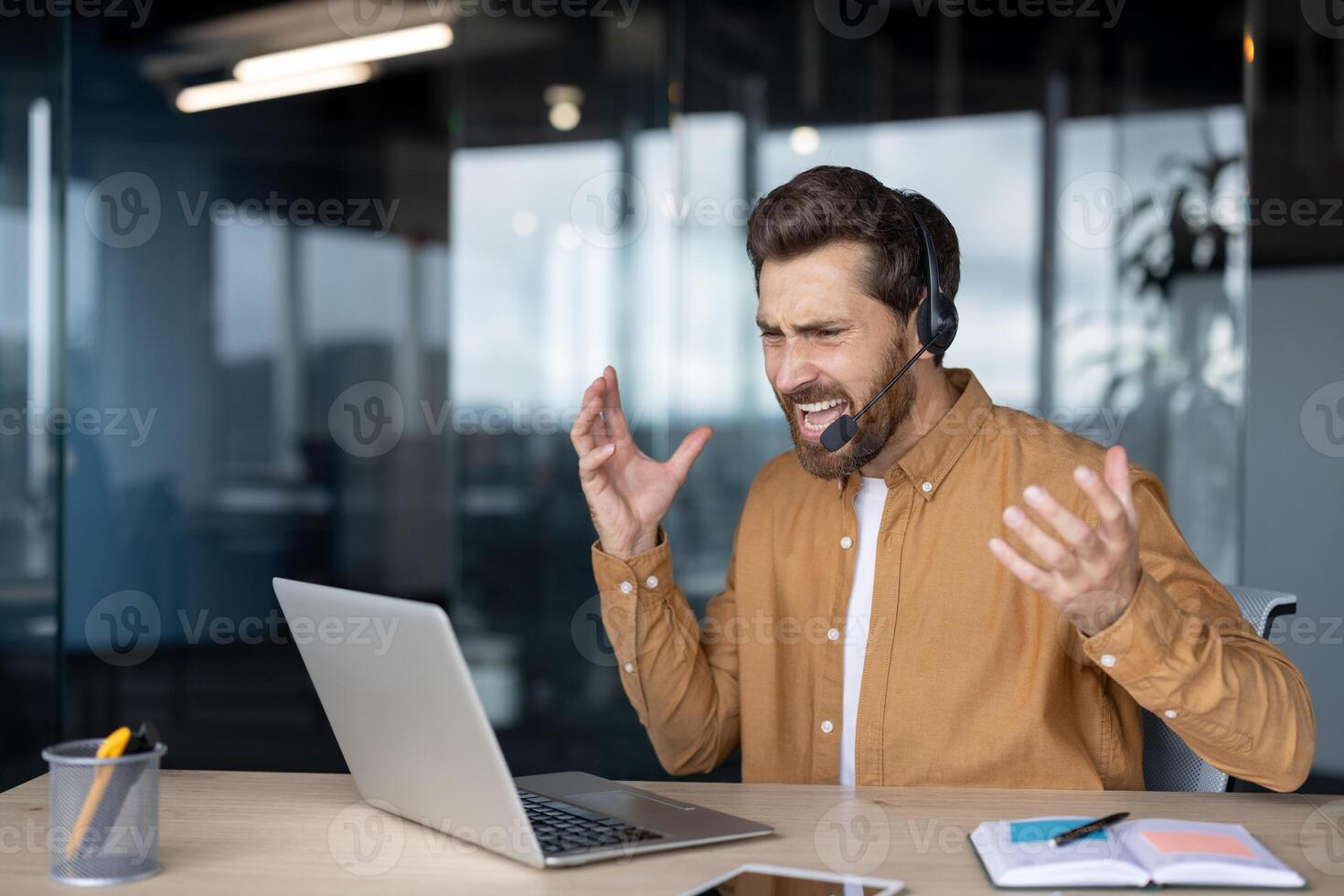 Businessman in modern office expresses frustration during call, using laptop with headset. Concepts include stress, technology issues, communication challenges. Ideal business emotional stress themes photo