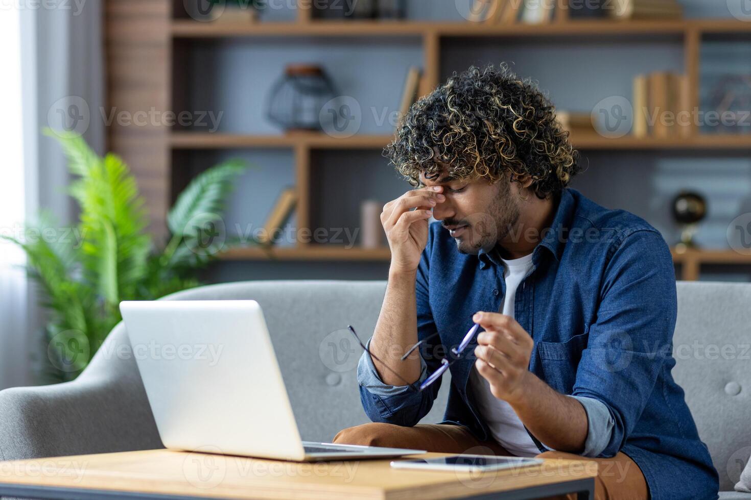 A tired man sits on a couch, rubbing his eyes, with a laptop and tablet nearby, suggesting stress or overwork. photo