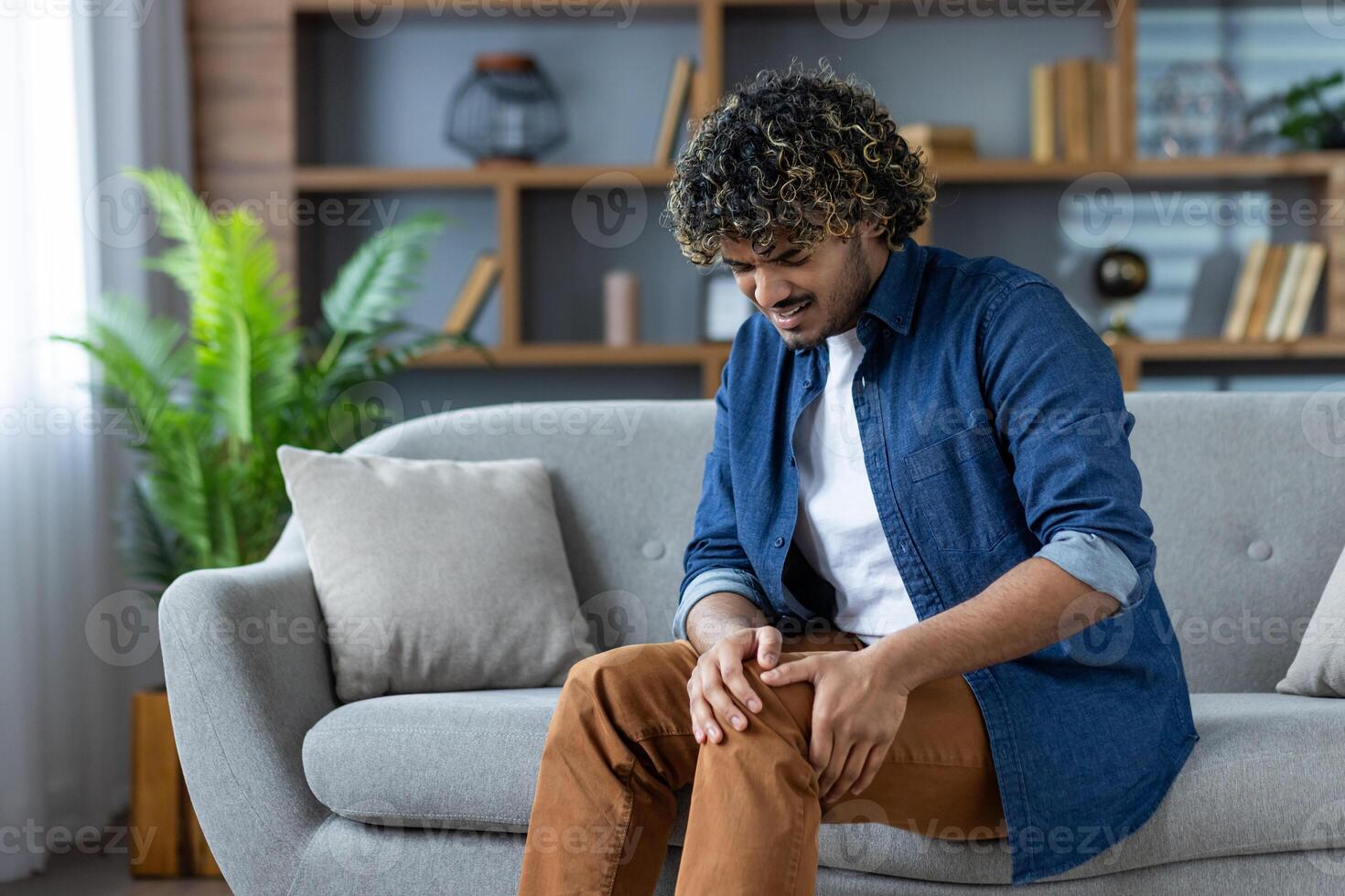 A man sitting on a couch clutches his knee in pain, indoors with shelves in the background. He appears to be experiencing discomfort. photo