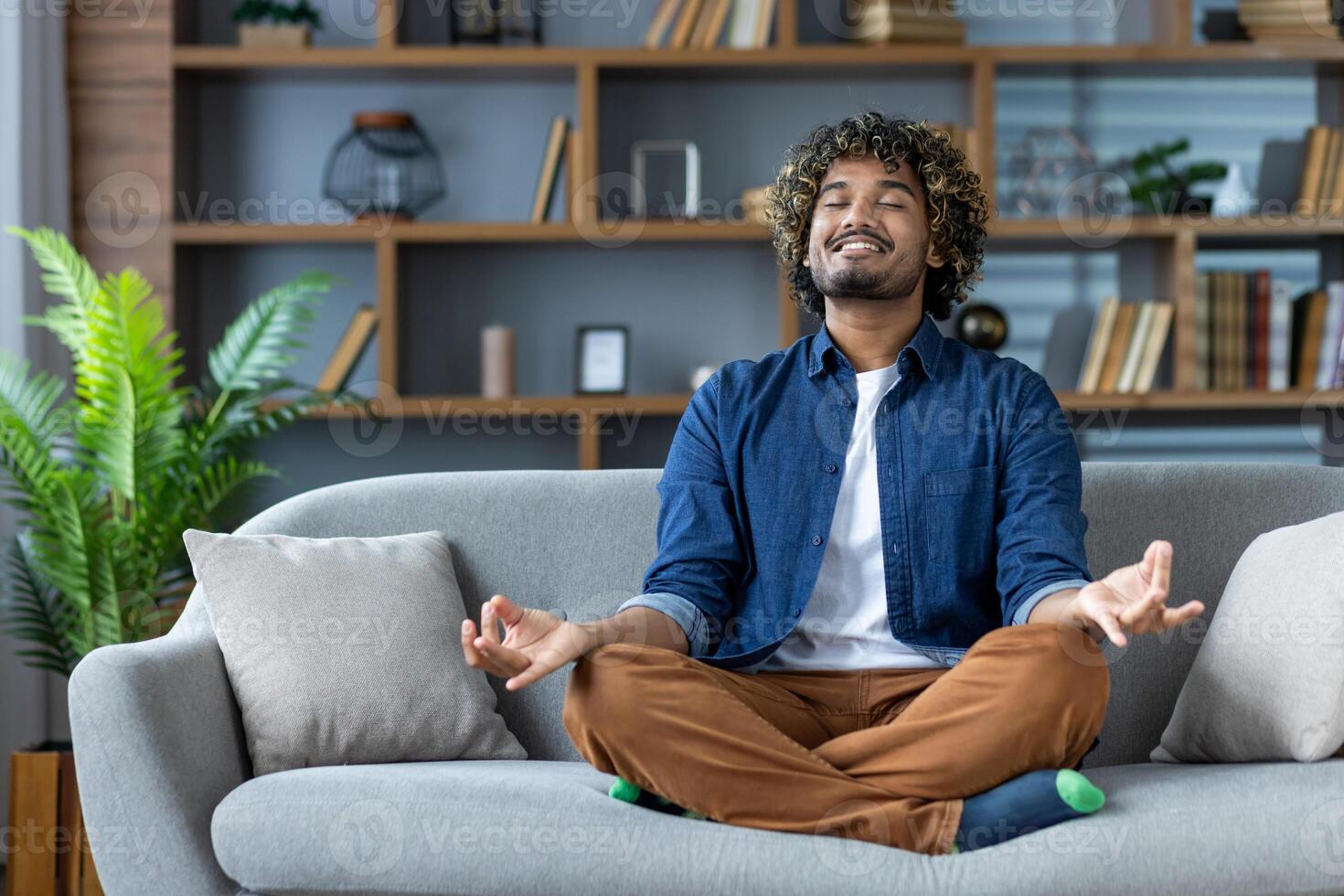 A man meditates peacefully at home, sitting comfortably on a sofa with his eyes closed and a calm expression. photo