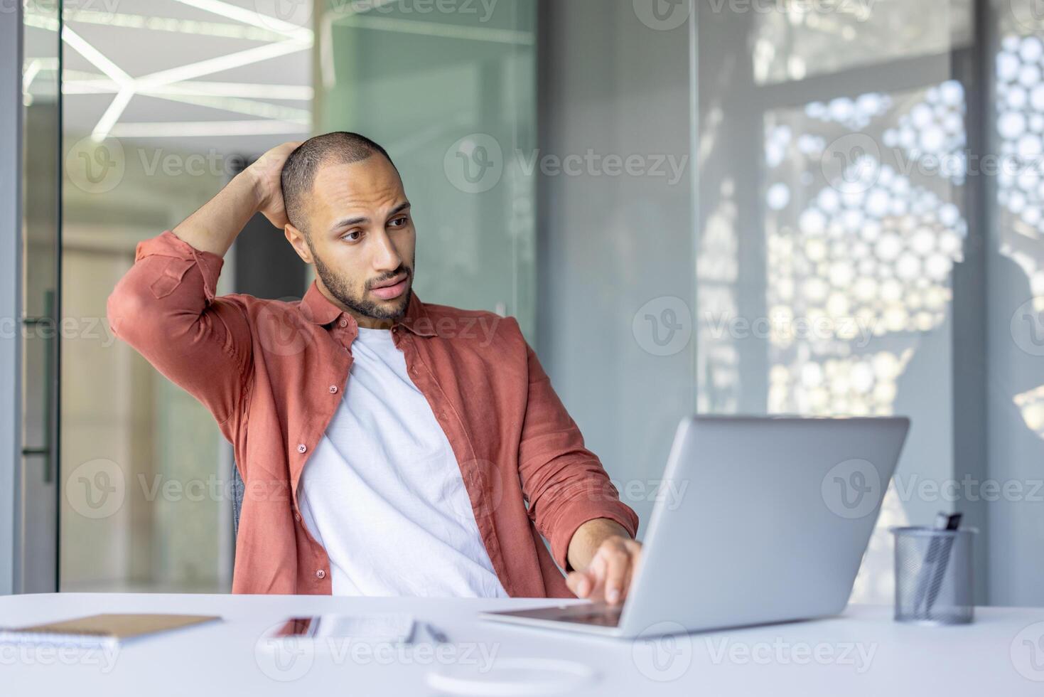 An African American man scratches his head looking thoughtfully at his laptop screen at a desk, facing some issue or idea. photo