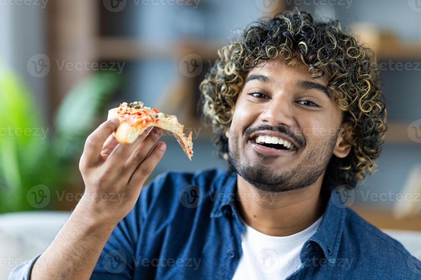 A smiling man with curly hair enjoys a slice of pizza, capturing the joy of a simple meal and a happy moment. photo