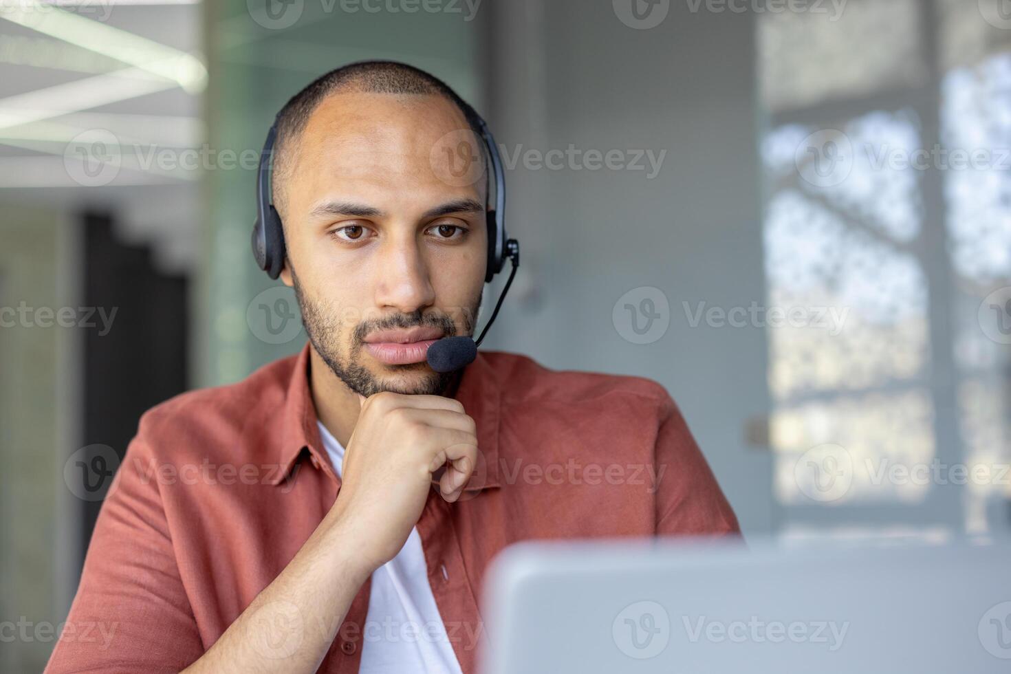 A thoughtful man wearing a headset, focused intently on his laptop screen. He's likely engaged in a virtual meeting or customer service call. photo