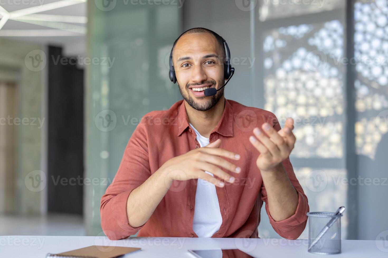 A smiling man wearing a headset gestures during a conference, sitting at a desk indoors, communicating. photo