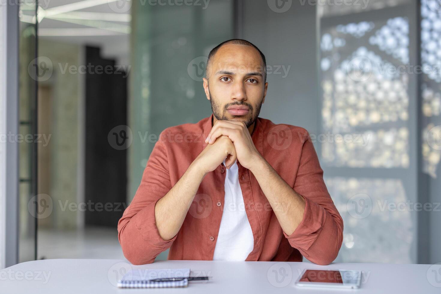 A serious man looks directly at the camera while sitting at a desk, appearing to be on a call photo