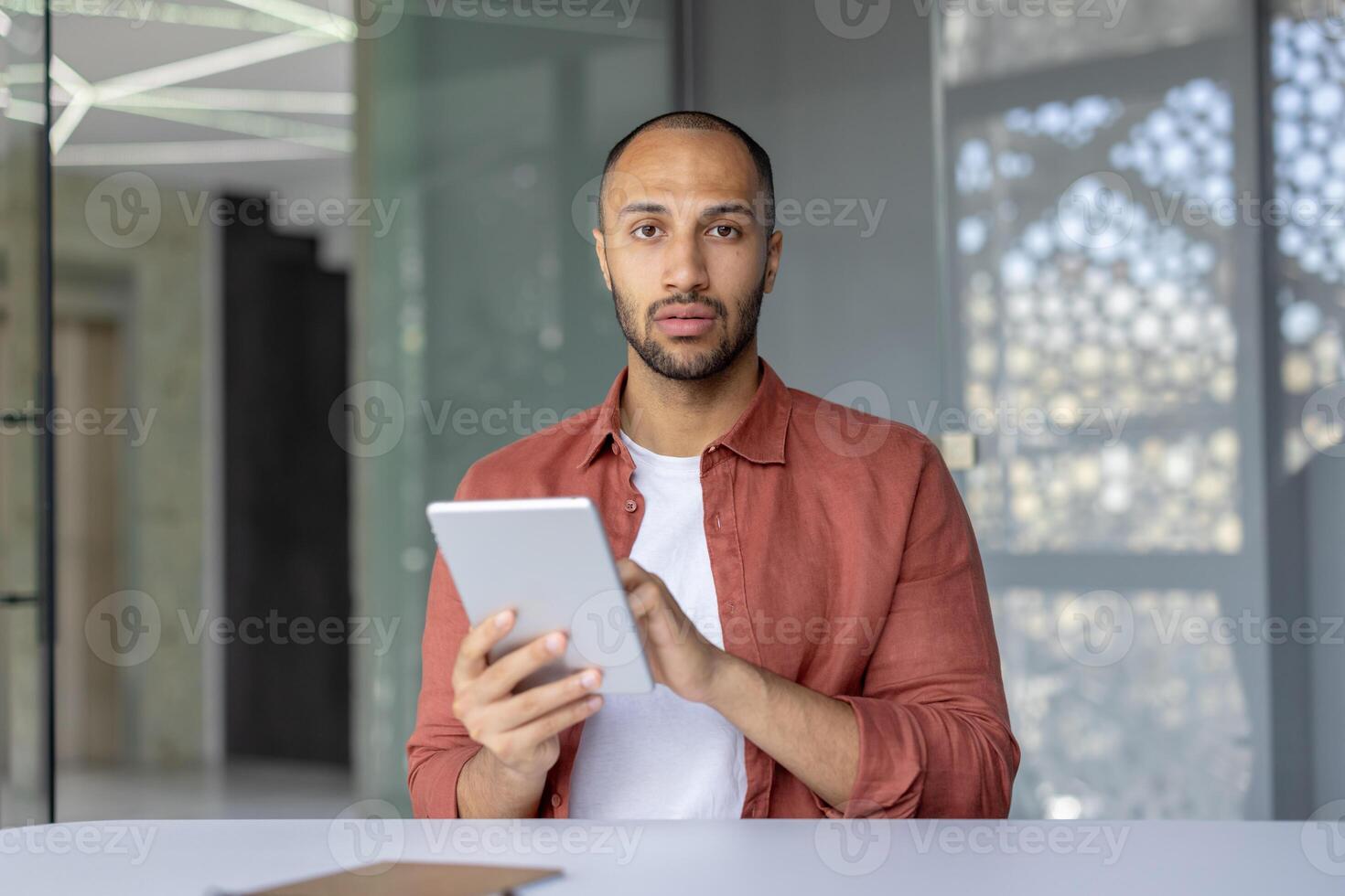 A man with dark hair and a red shirt is looking at the camera while holding a tablet. photo