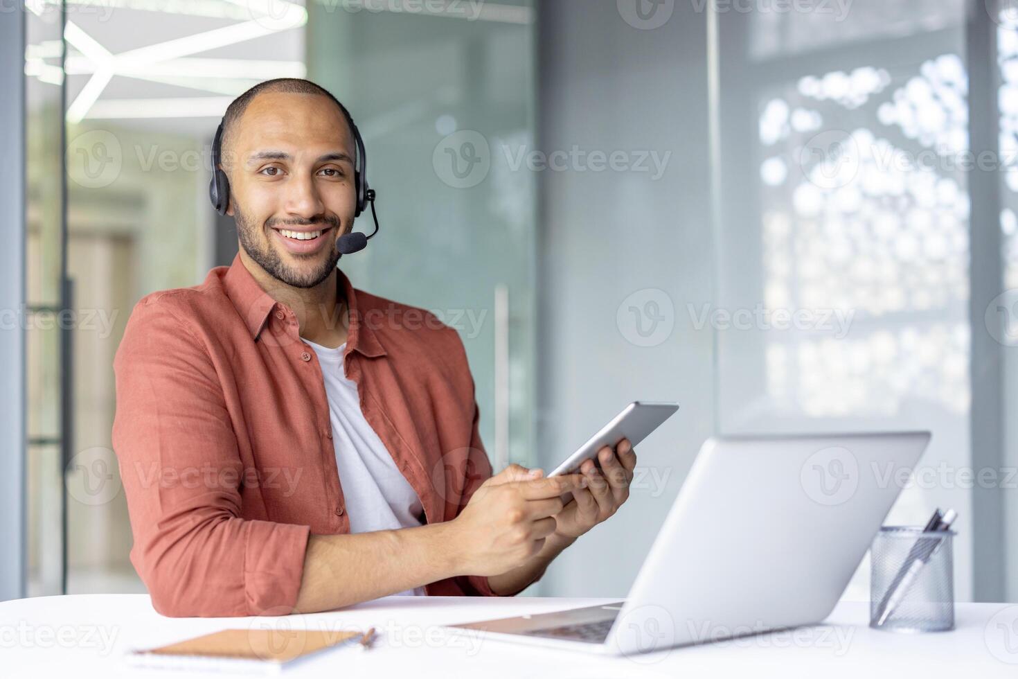 A smiling man wearing a headset, holding a tablet, and sitting at a desk in an office setting, using technology. photo