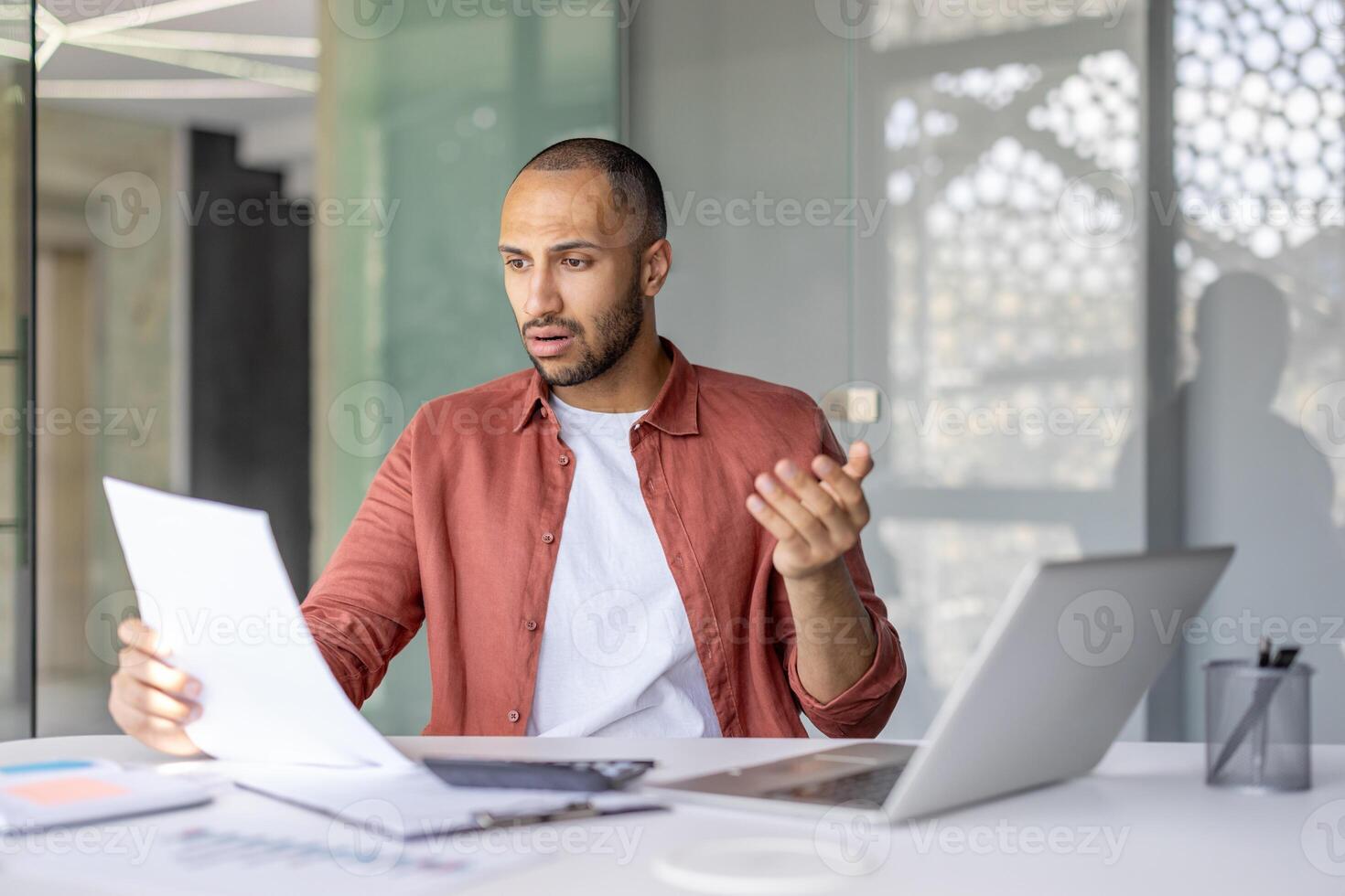 A surprised man examines documents while sitting at a desk. A laptop and office supplies are present, too. He seems bewildered by the information. photo