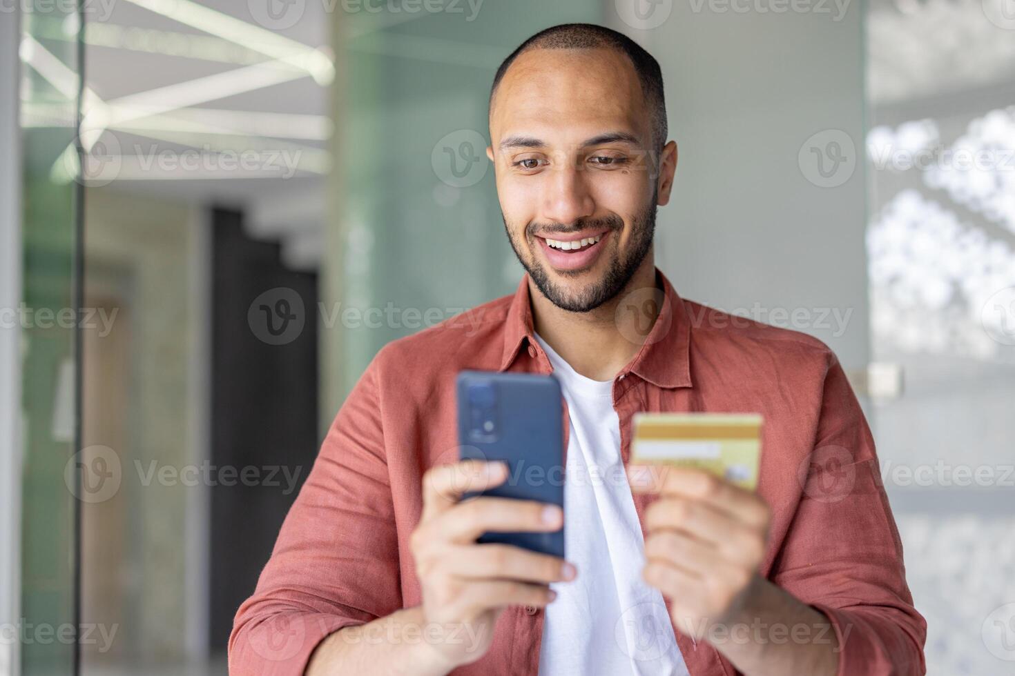 Close-up photo, man with phone and bank credit card smiling makes online purchases in online store remotely. Businessman inside office at workplace transfers money. photo