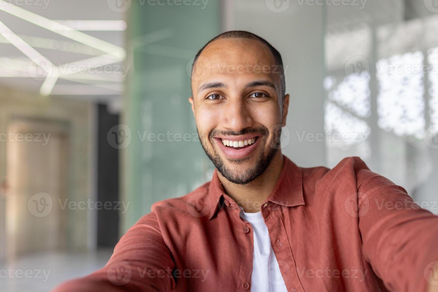 A close-up shot captures a smiling man in a casual orange shirt, taking a selfie indoors with a bright, welcoming expression. photo