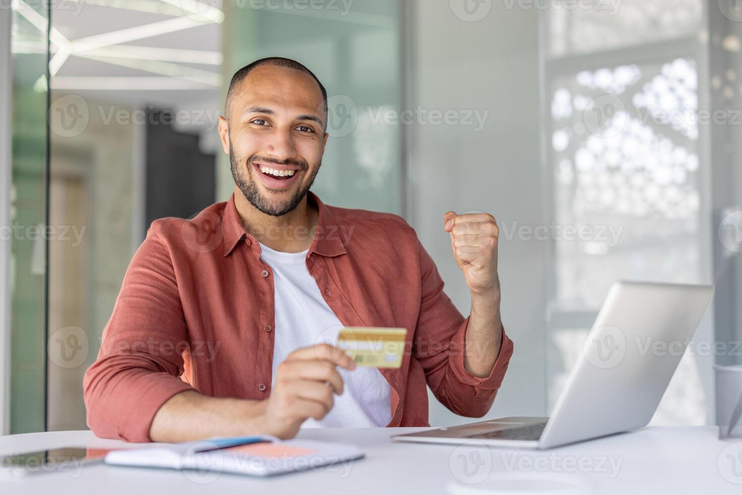 Joyful successful man with bank credit card and laptop looking at camera. Company employee celebrating successful online money transfer. photo