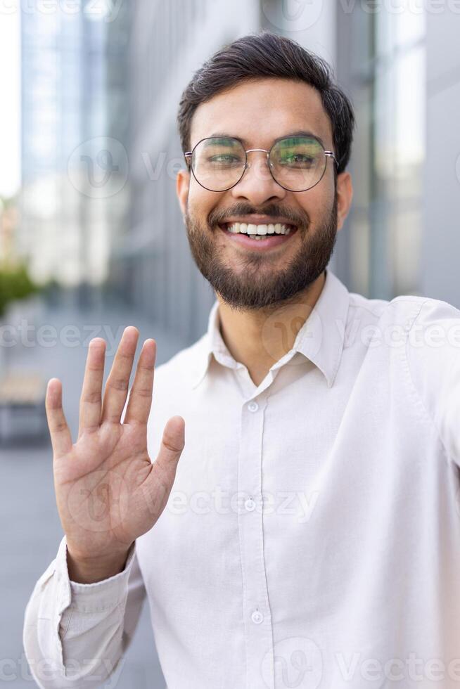 A man with a beard and glasses waves during a call outside a building photo