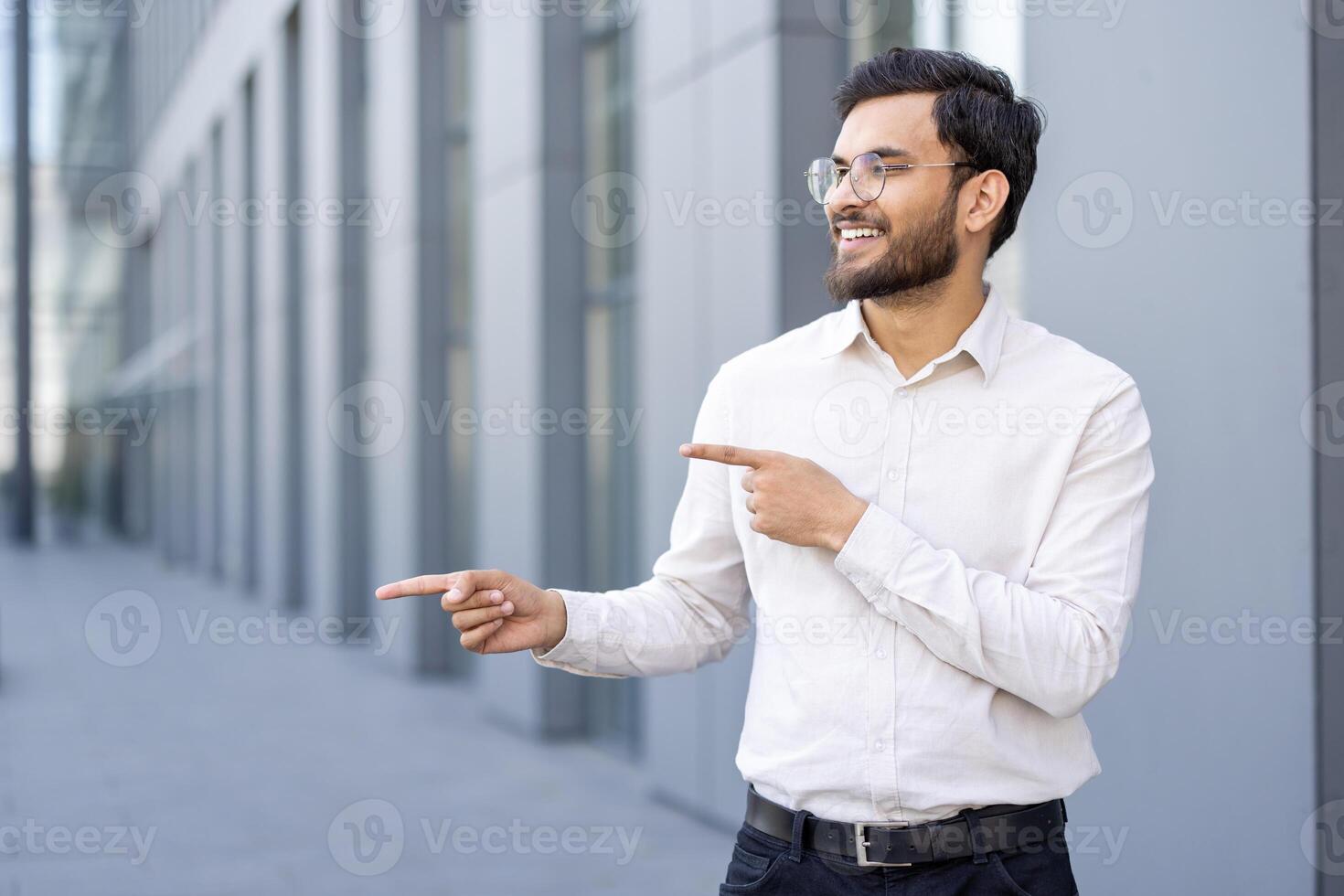 A smiling businessman of Indian descent points sideways, wearing glasses and a button-down shirt, outdoors with a modern building in the background. photo