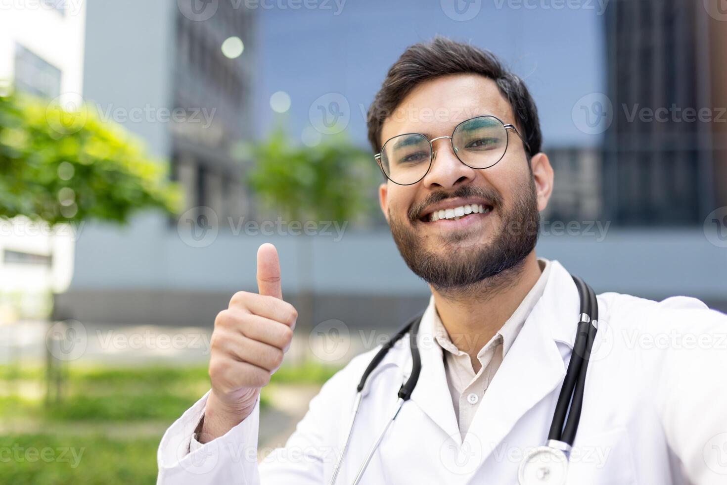 A smiling doctor wearing glasses gives a thumbs up outside, looking positive and optimistic. He is wearing a white lab coat with a stethoscope around his neck. photo