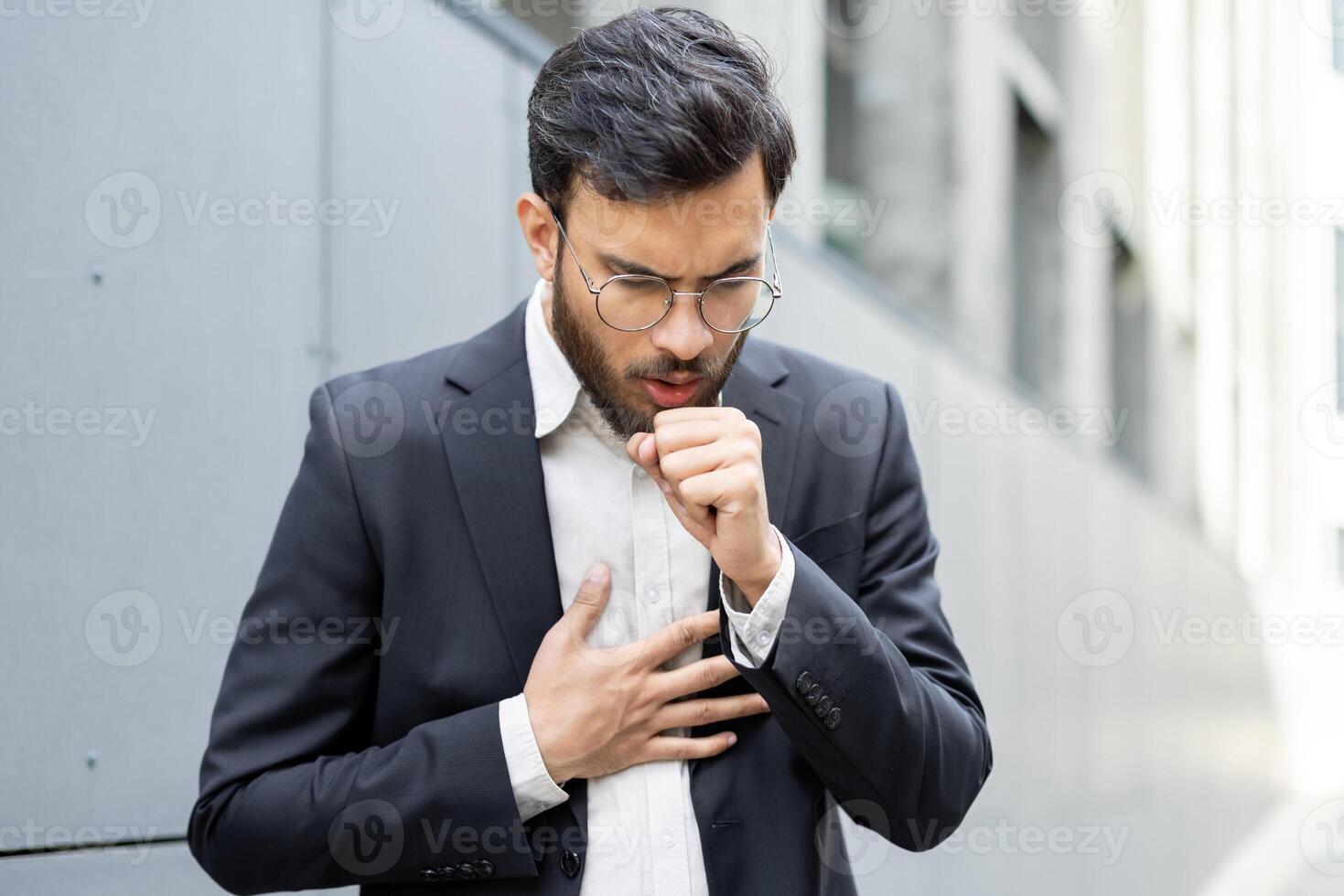 A man in a suit is coughing outdoors, looking unwell. He is holding his chest, likely experiencing discomfort or illness. photo