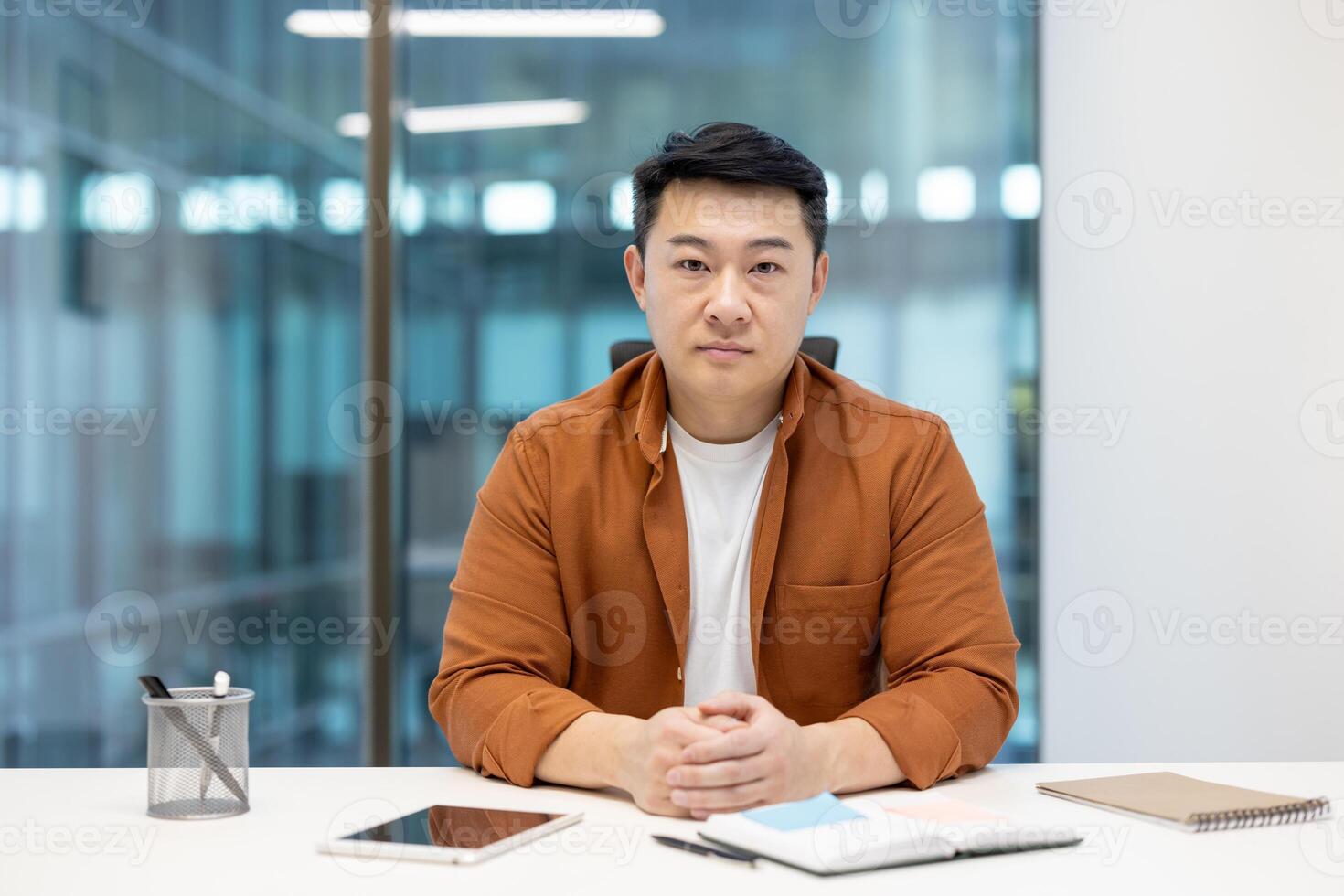 A focused Asian man sits at a desk, looking directly at the camera in a modern office setting, wearing a button-down shirt. photo