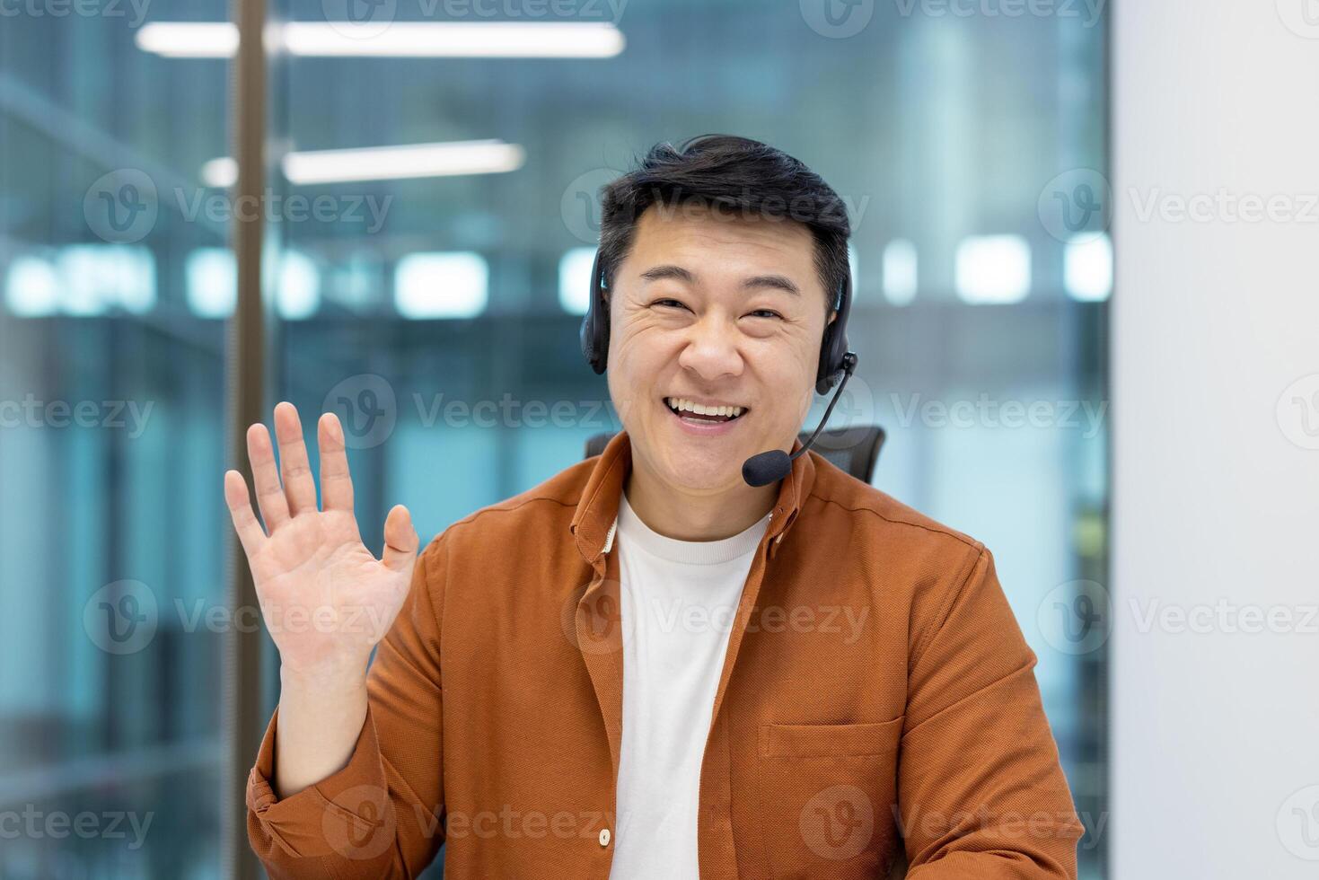 A cheerful Asian man wearing a headset waves at the camera, smiling warmly, in a bright office setting. photo