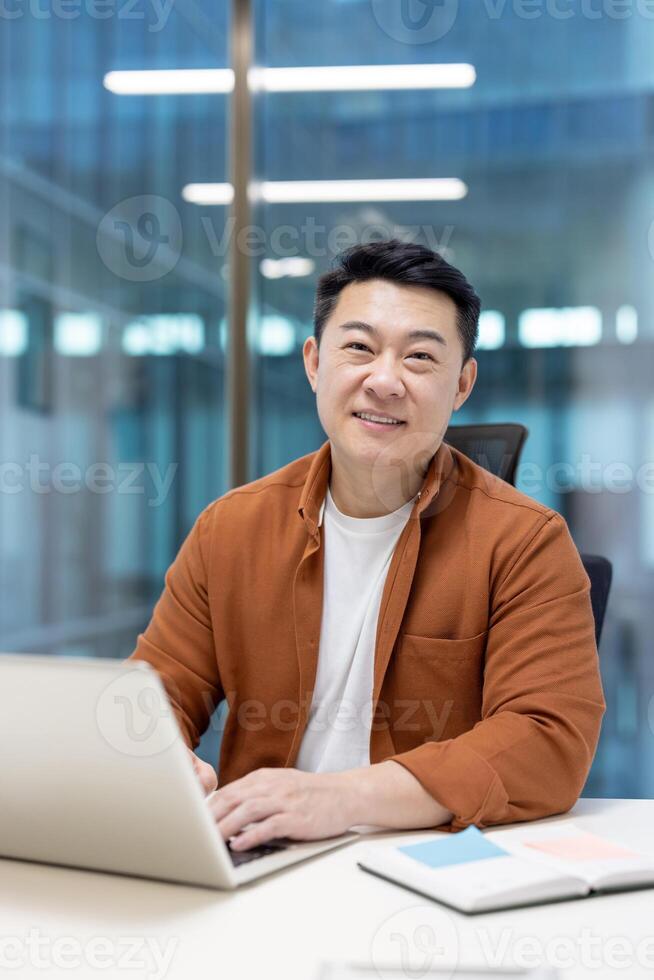 An Asian man smiling while working on a laptop in a modern office environment. photo