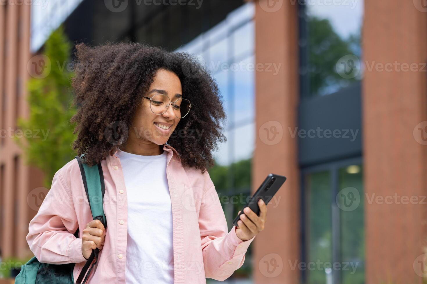 A smiling woman holds a phone and looks at it outdoors. She wears glasses and a backpack, and is walking in front of a building. photo