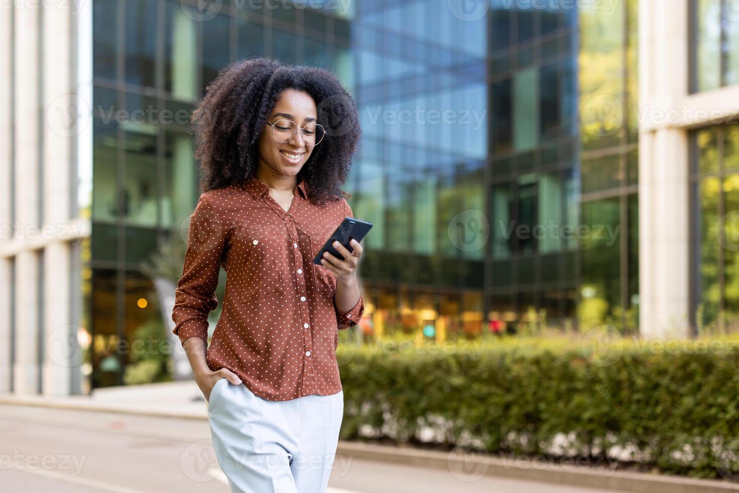 A smiling woman in glasses uses her phone while walking outside near a modern office building. photo