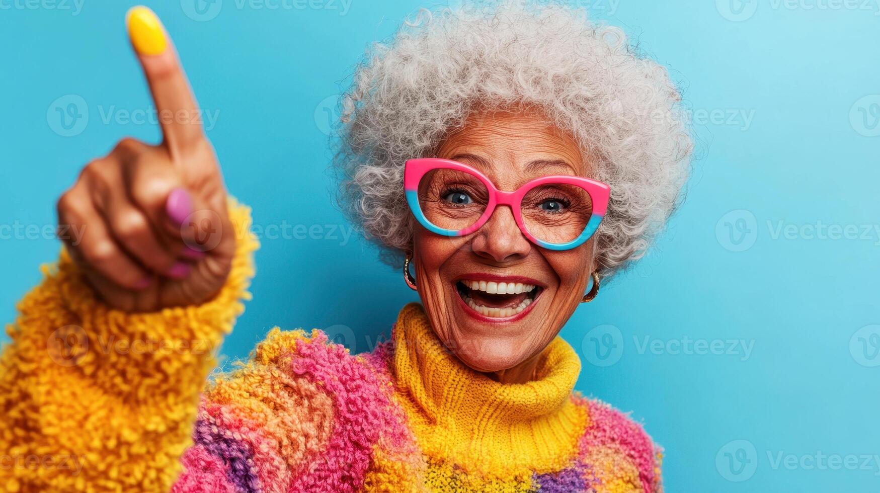 Joyful older woman with colorful sweater and glasses pointing against a bright blue background in a cheerful expression celebrating life and positivity photo