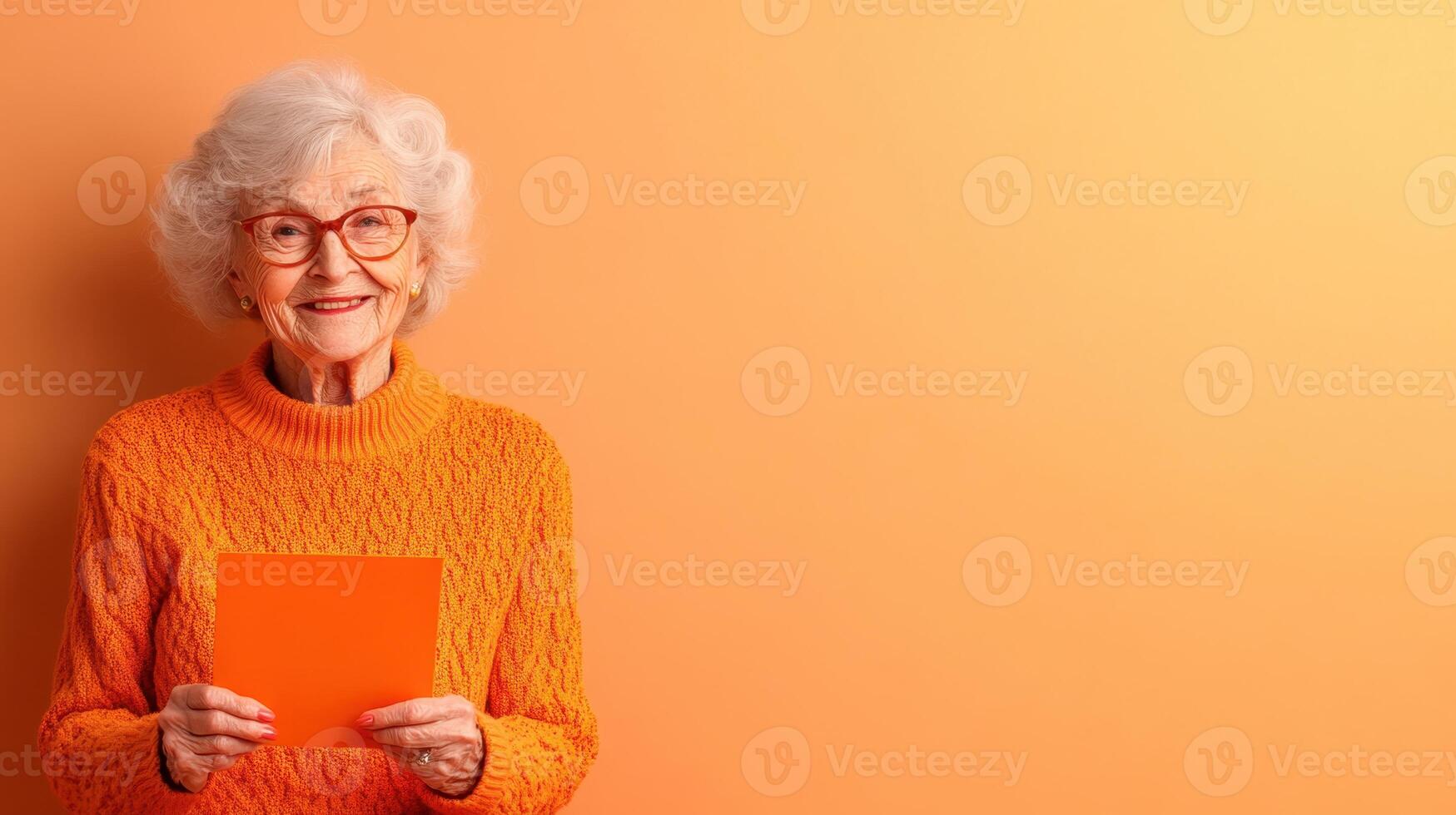 Elderly woman in orange sweater smiling while holding a bright orange card against a soft orange background in a cheerful setting photo