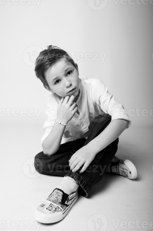 Black and white portrait of a young boy lying on his stomach, chin resting on hands, looking at the camera with a dreamy, thoughtful expression. Soft shadows create a calm mood. photo