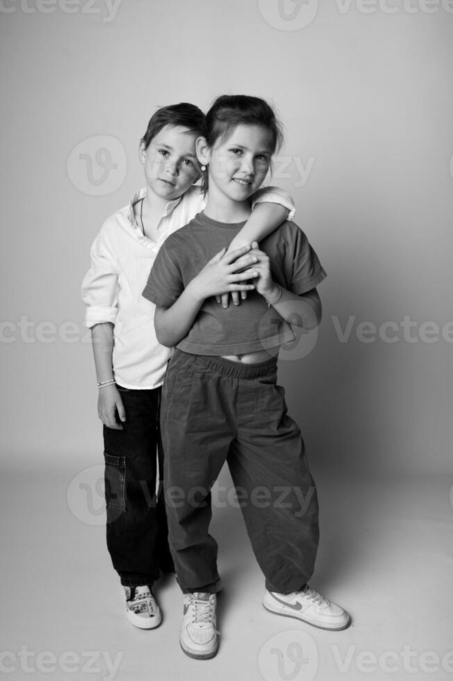 Monochrome full-length photo of a boy and girl standing slightly apart, looking at the camera with serious faces. Strong contrast and minimalist backdrop.